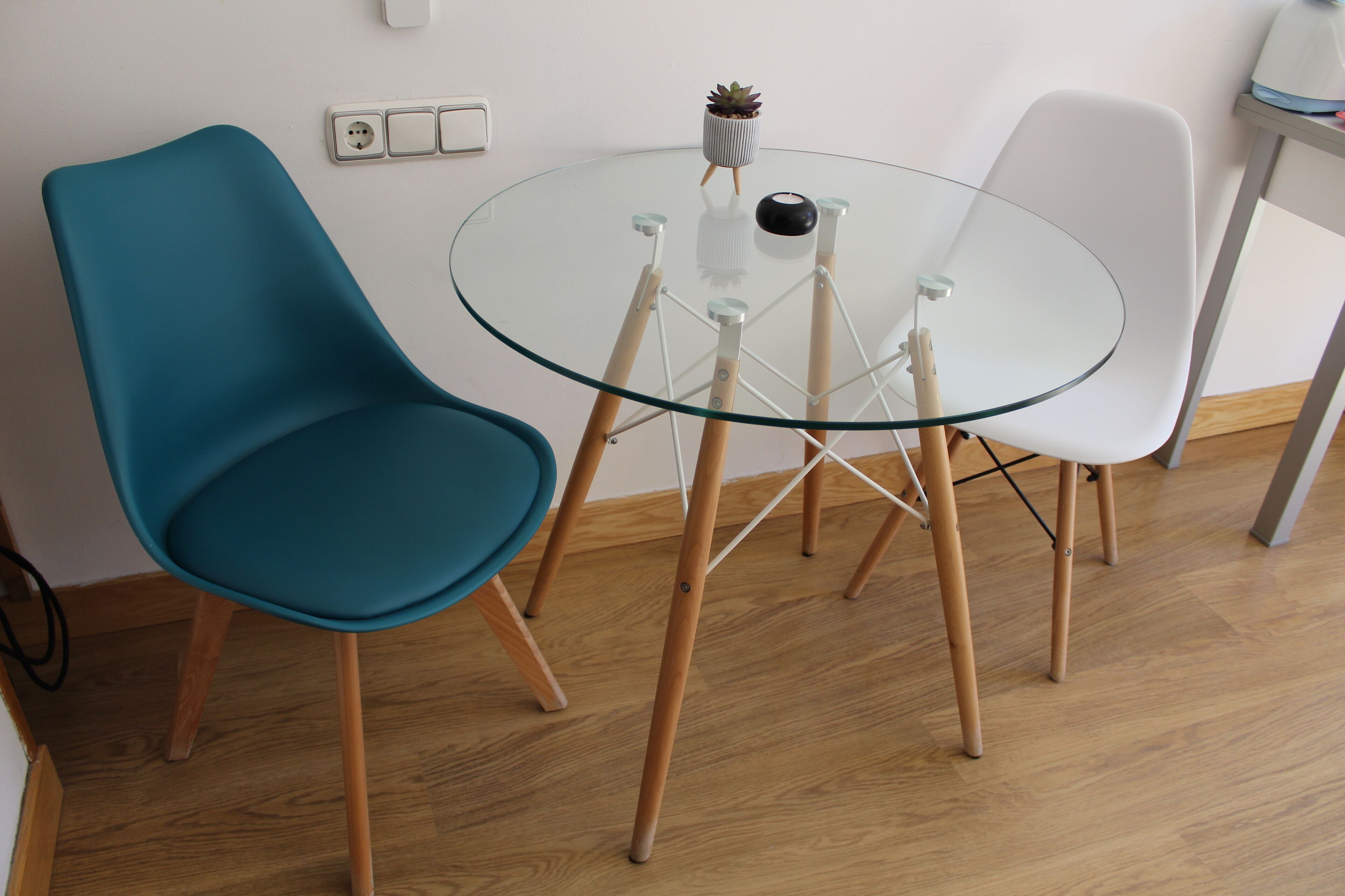 A room with a glass table surrounded by four chairs, a small potted plant, and a power outlet on the wall.