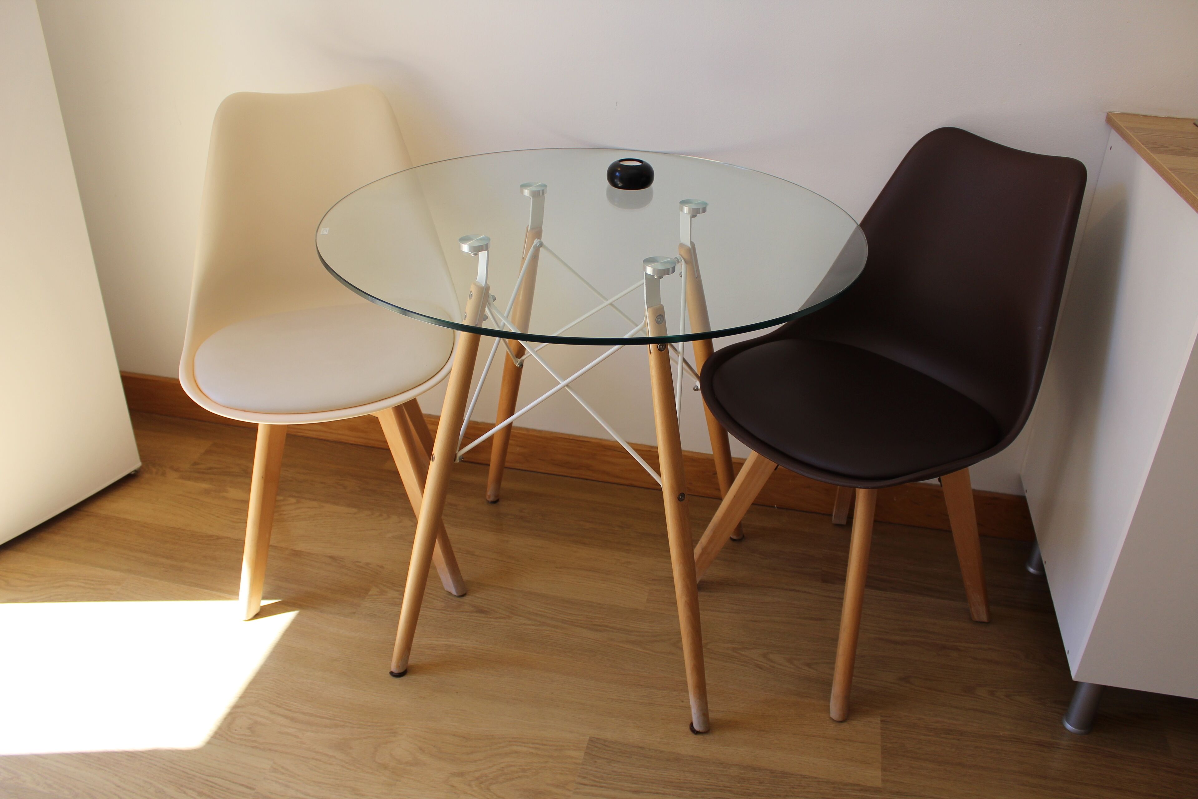 A room with a round glass table and two chairs, one beige and one black, with wooden legs.