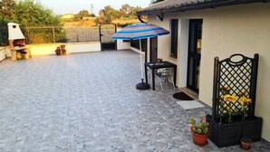 Un espace patio doté d’un sol carrelé, d’un barbecue, d’une table avec des chaises et d’un parasol à rayures.