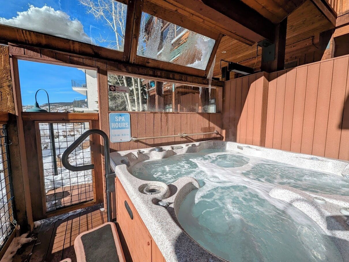 A hot tub with a view of a snowy landscape, wooden walls, and a sign indicating spa hours.
