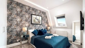 A bedroom with a large bed, a nightstand, a lamp, a framed picture on the wall, and a window with a view.
