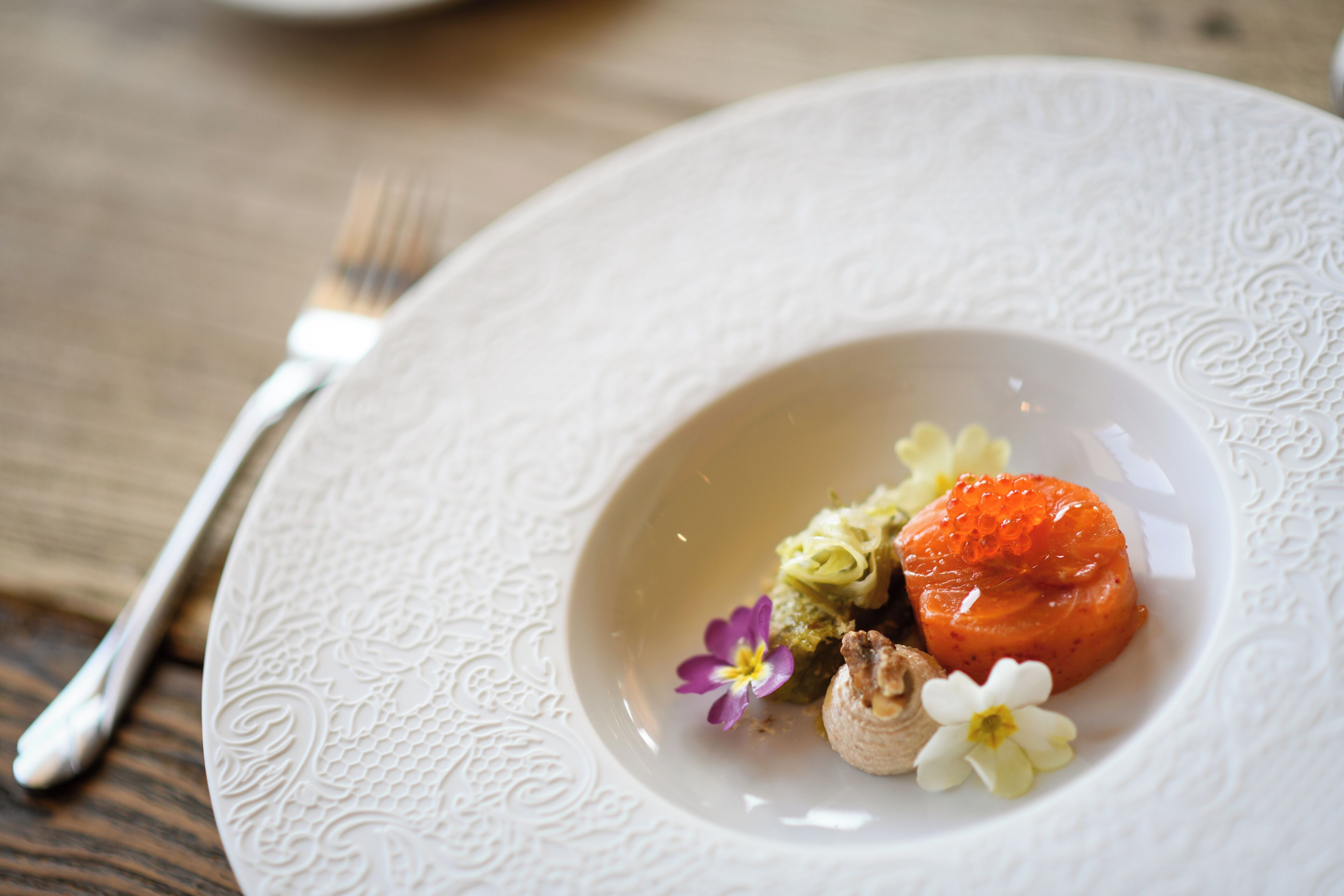 A white plate with intricate patterns, containing a salmon tartare, a small flower, and a mushroom.