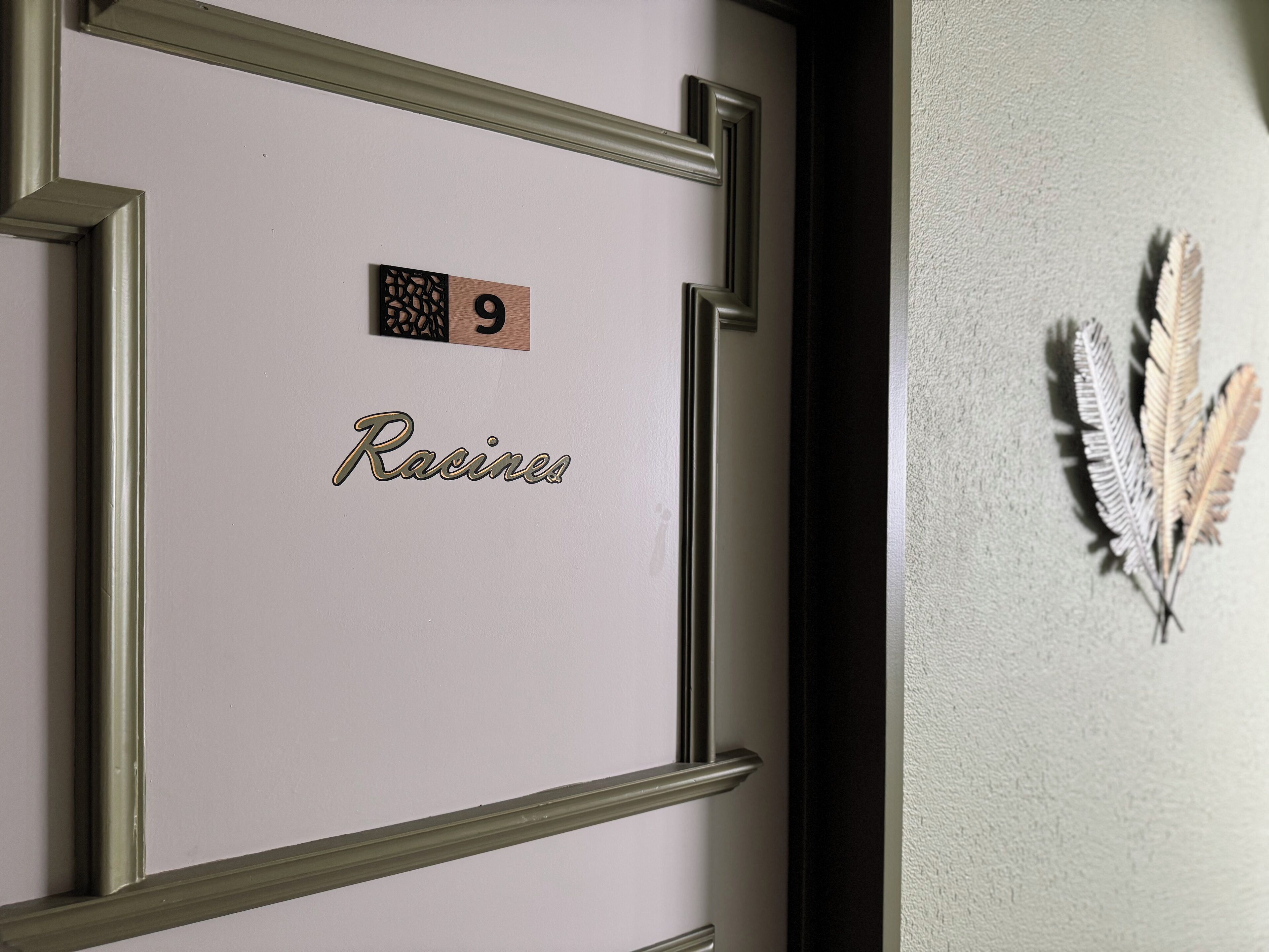 Une porte portant le numéro 9 et le mot « Racines », une plume décorative accrochée au mur et un cadre de porte orné d’un motif géométrique.