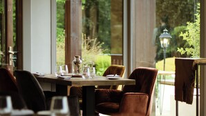 Un coin repas avec une table dressée pour deux personnes, de grandes fenêtres et une vue sur la verdure environnante.