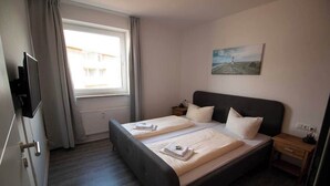 A bedroom with two beds, a TV, a painting, and a window with curtains. - Hafen Apartments  (Aurich)