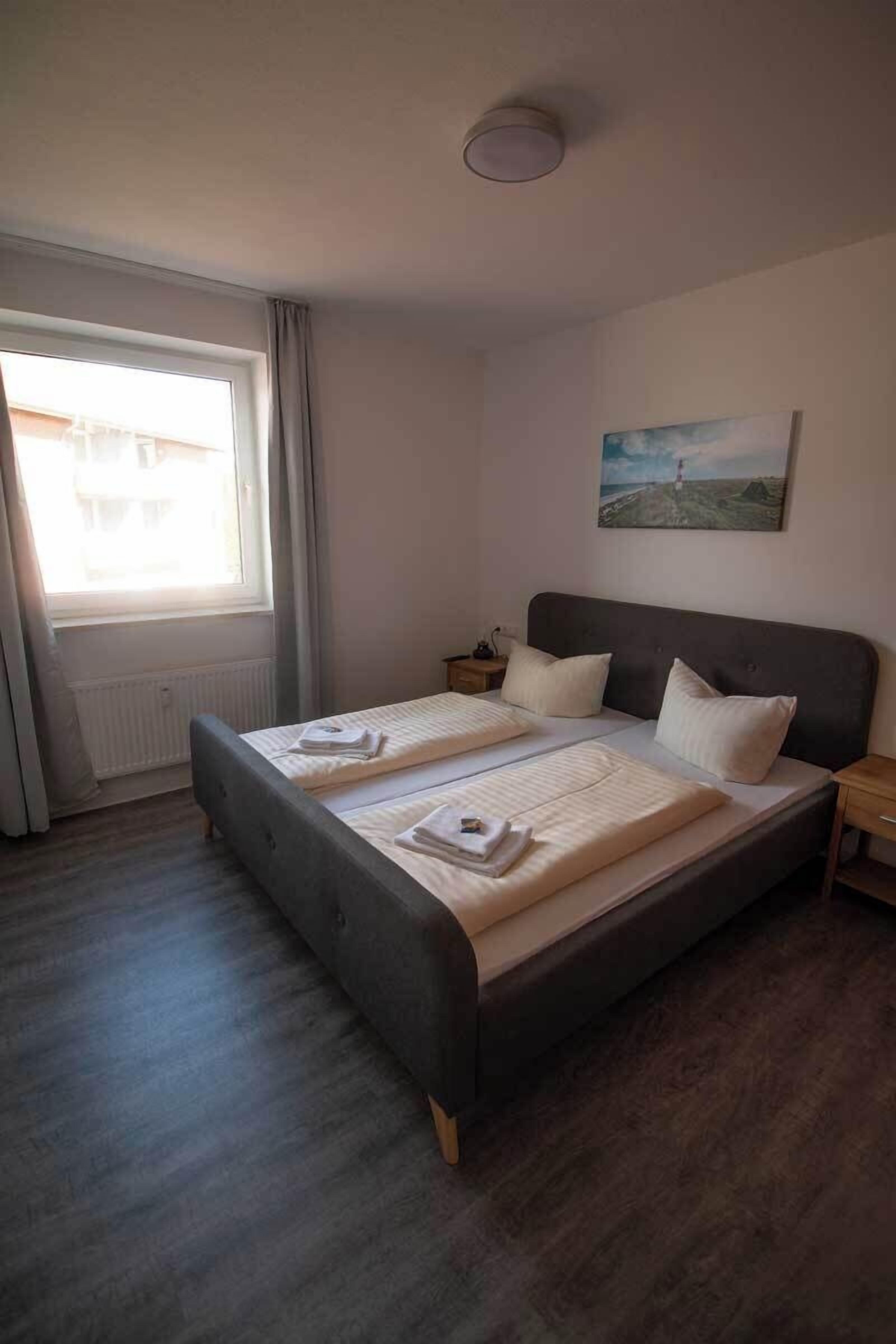 A bedroom with a large bed, two bedside tables, a window with curtains, and a painting on the wall.