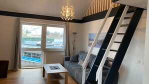 A modern living room with a sofa, a coffee table, a staircase, and a large window with a view of a construction site. - Hafen Apartments  (Aurich)
