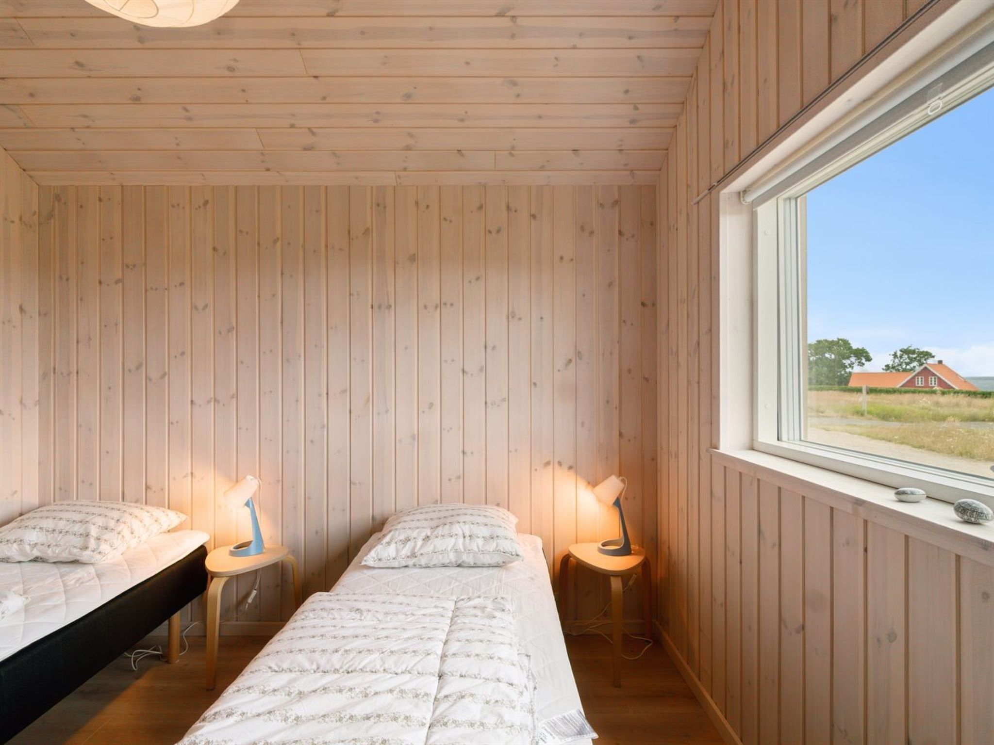 Ein Zimmer mit Holzwänden und einem großen Fenster. Es gibt zwei Betten mit weißer Bettwäsche sowie zwei Nachttische mit Lampen.