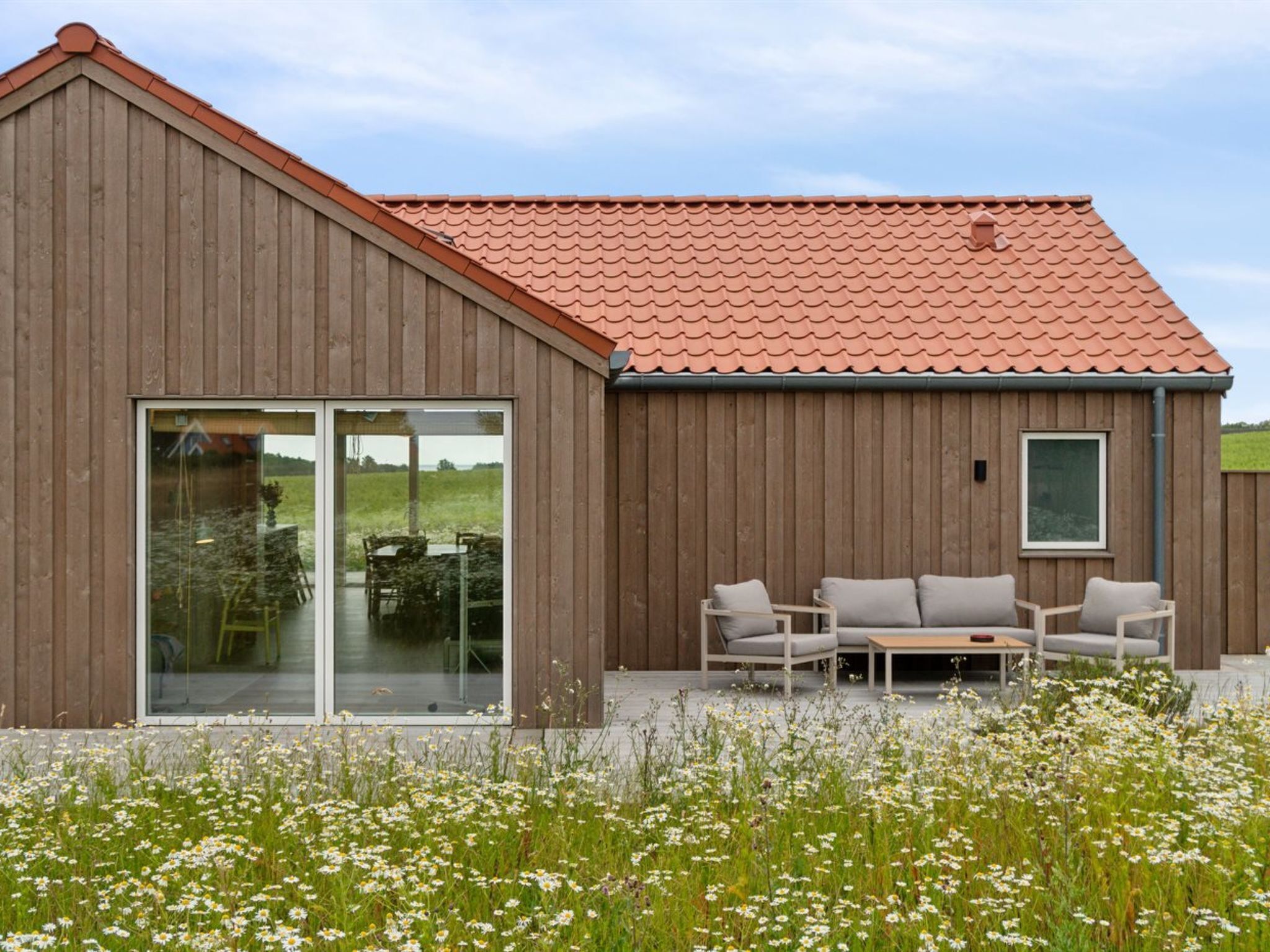 A modern wooden building with a red-tiled roof, large glass windows, and a patio area with outdoor furniture.
