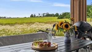 Une table en bois sur laquelle se trouvent un plateau de fruits et de boissons, un barbecue, et d’où l’on peut admirer un champ de tournesols.