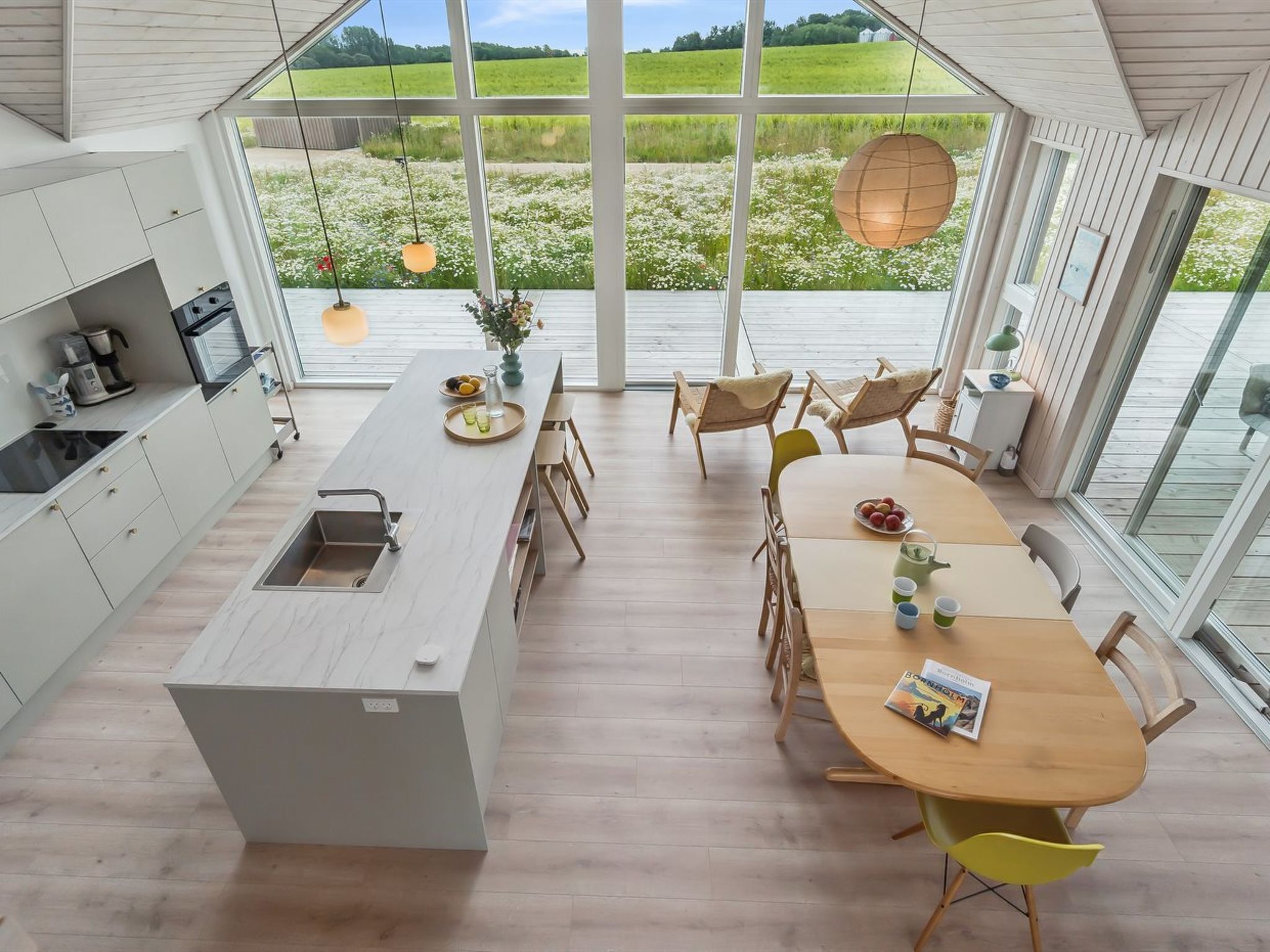 A modern kitchen with a large island, wooden dining table, and chairs. There is a view of a green field through large windows.