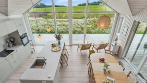 Une cuisine moderne dotée d’un grand îlot central, d’une table à manger en bois et de chaises. De grandes fenêtres offrent une vue sur un champ verdoyant.