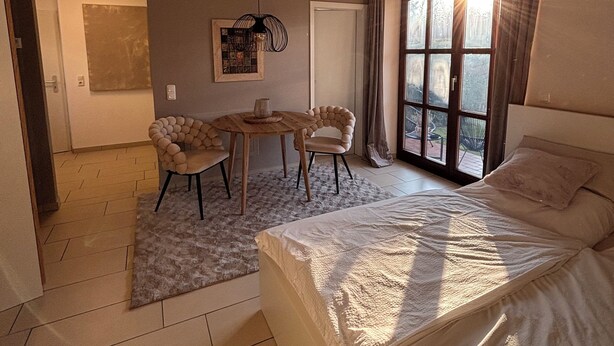 A bedroom with a bed, a desk, two chairs, a hanging light fixture, and a window with sunlight.