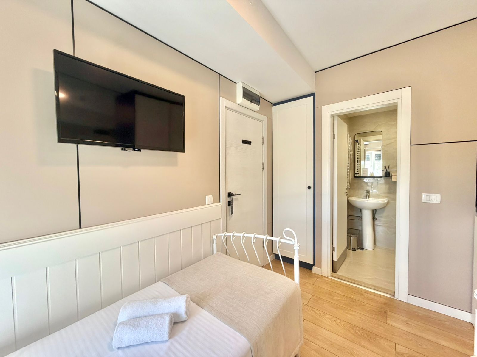A bedroom with a bed, a TV mounted on the wall, a door leading to a bathroom, and a wooden floor.