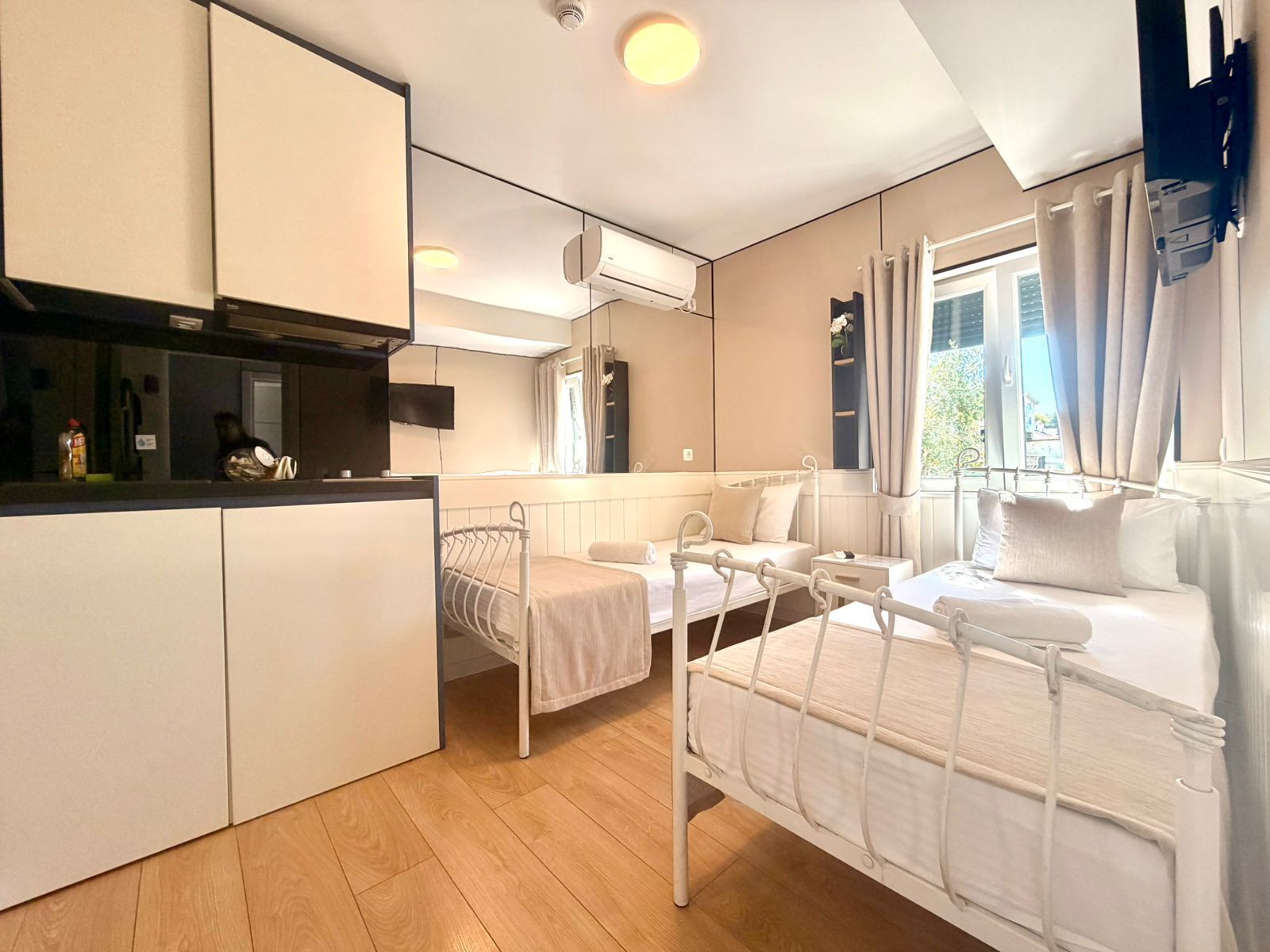 Habitación de hotel pequeña con kitchenette, dos camas individuales, un espejo grande y ventana con cortinas.