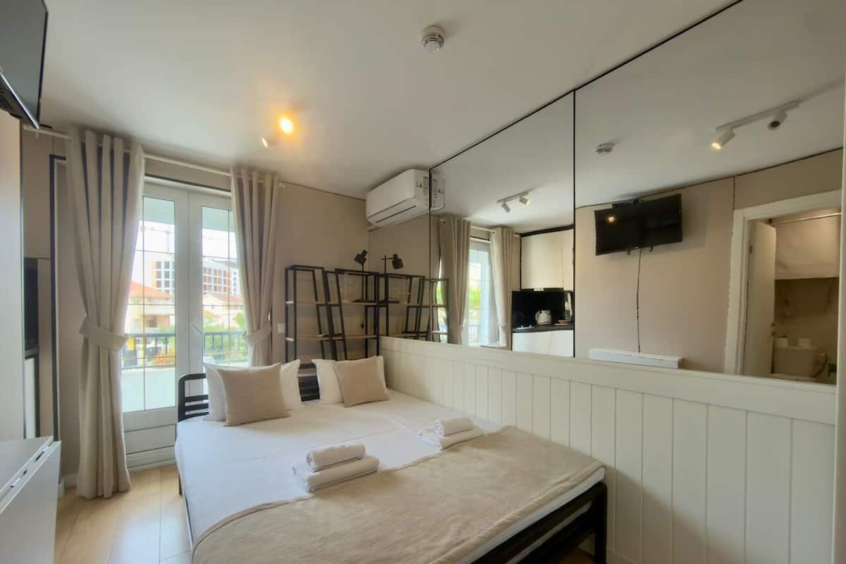 A modern hotel room with a large bed, a flat-screen TV, an air conditioning unit, and a view of the city through the window.