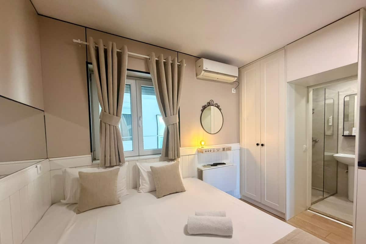 A bedroom with a bed, curtains, an air conditioning unit, a mirror, a bedside table, and a bathroom with a shower.
