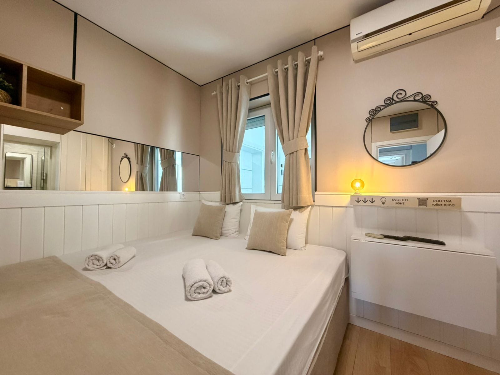 Une chambre d’hôtel comprenant un lit, un miroir, une fenêtre avec des rideaux, un climatiseur fixé au mur et un panneau de commande permettant de régler l’éclairage et les stores.