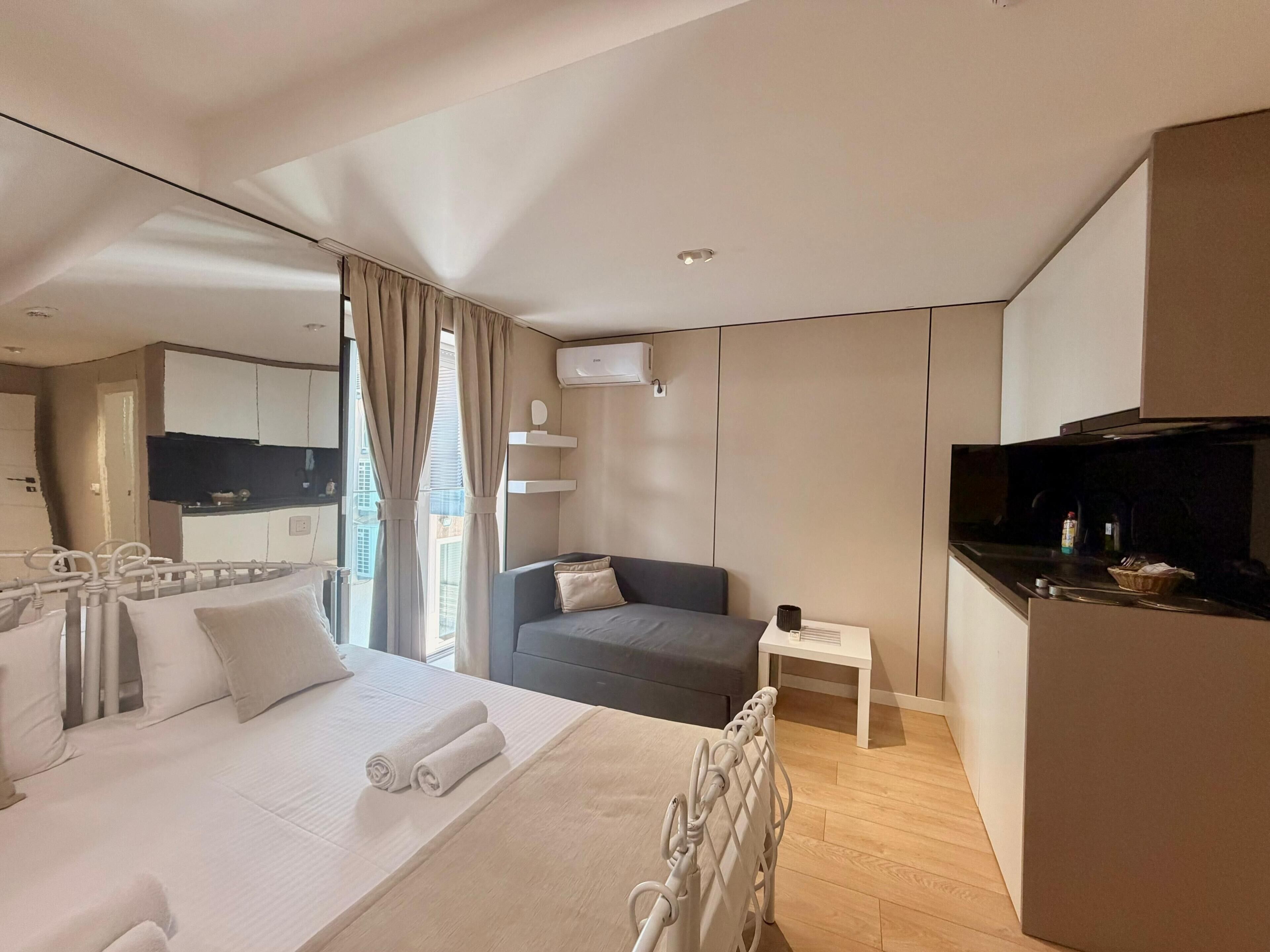 Quarto de hotel moderno com cama, sofá, mesa pequena e cozinha compacta.