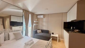 Quarto de hotel moderno com cama, sofá, mesa pequena e cozinha compacta.