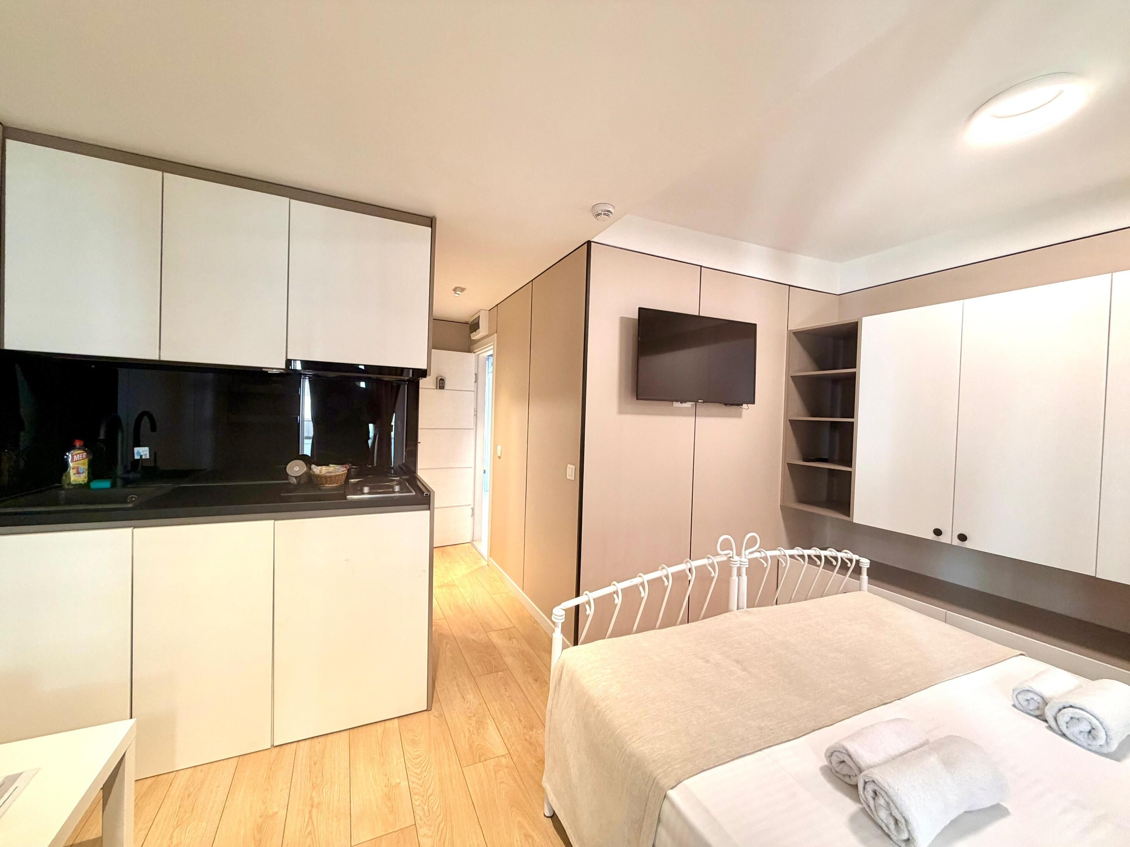 A compact hotel room with a kitchenette, a bed with towels, and a TV mounted on the wall.