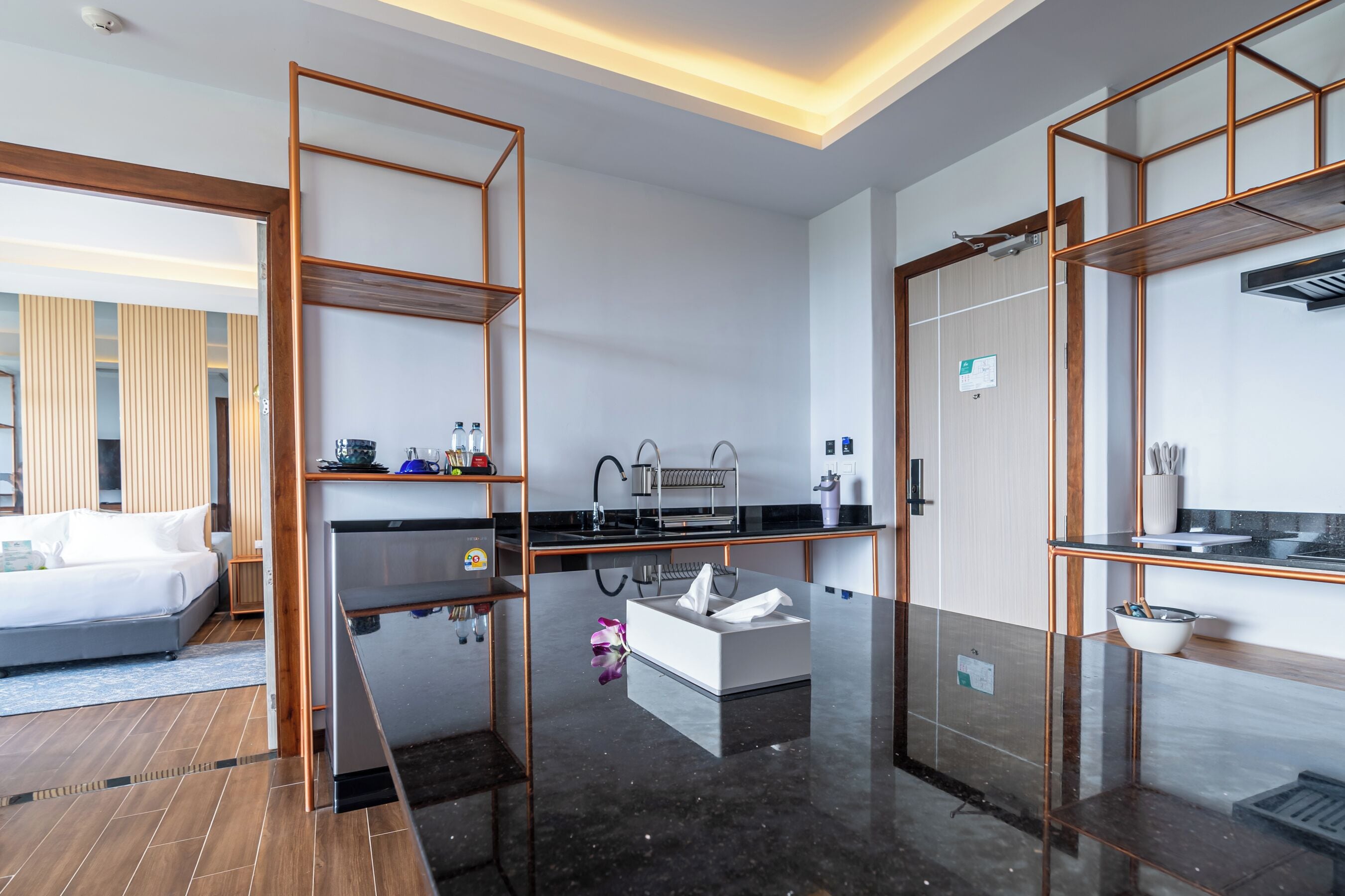A modern hotel room with a large bed, a kitchen area with a sink and cabinets, and a bathroom with a mirror and towel rack.
