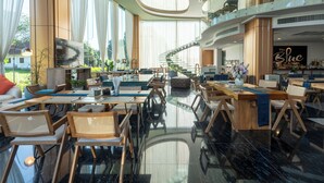 A spacious hotel lobby with a modern design, featuring a central seating area with tables and chairs, a staircase leading to an upper level, and large windows allowing natural light. - The Blue Mavi Hotel (Chalong)