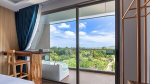 A modern hotel room with a large sliding glass door leading to a balcony, a wooden desk, a comfortable chair, and a bed. - The Blue Mavi Hotel (Chalong)