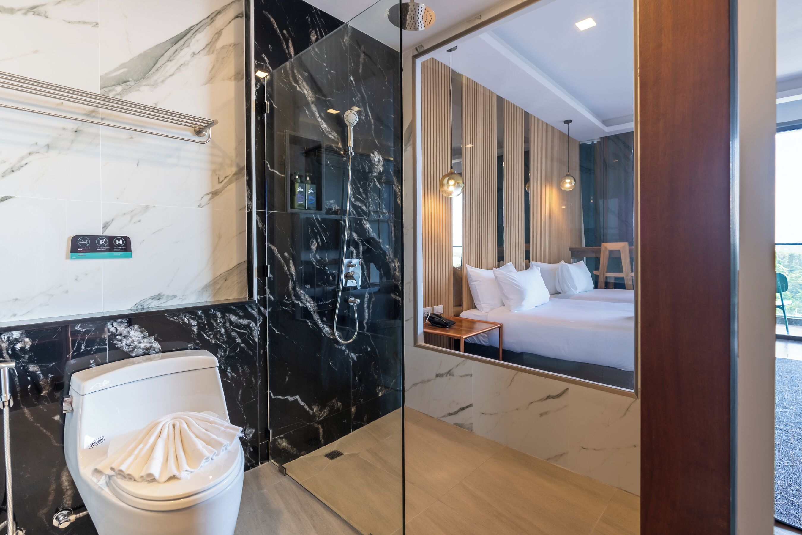 A modern bathroom with a marble shower, a toilet, and a mirror reflecting a bedroom with a bed and a nightstand.
