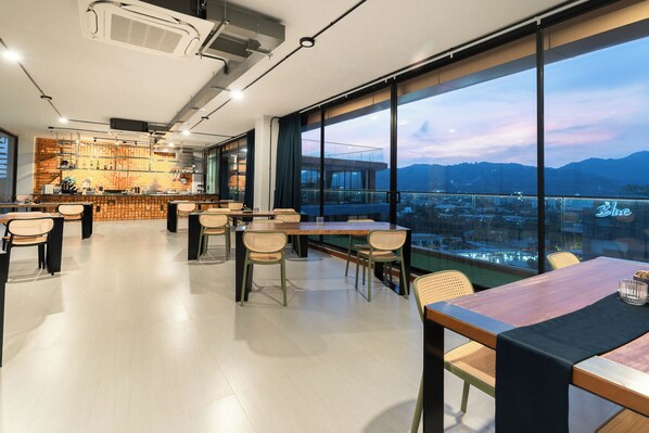 A modern restaurant with a bar area, large windows offering a city view, and a mix of wooden and metal furniture. - The Blue Mavi Hotel (Chalong)