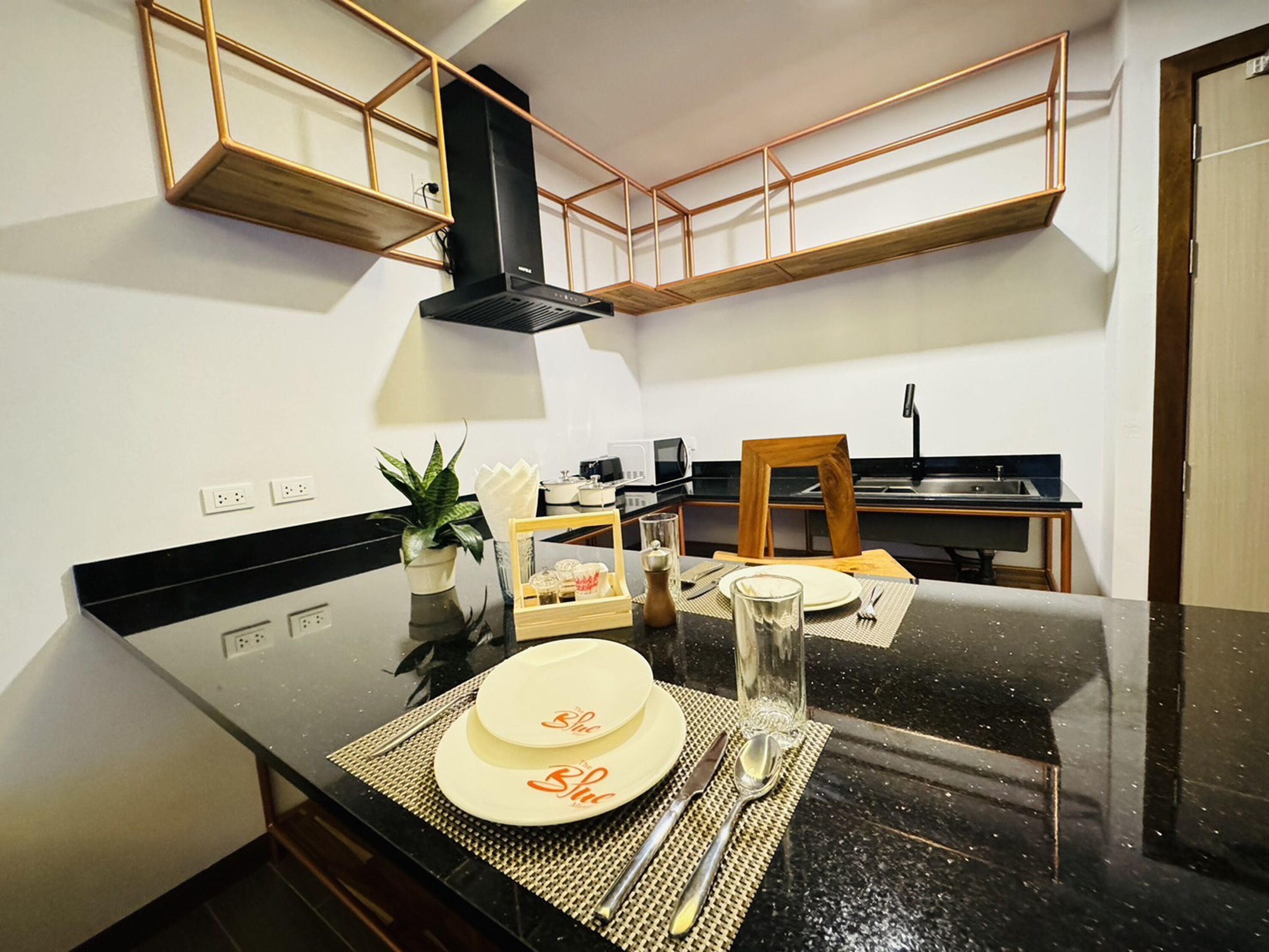 A modern kitchen with black countertops, wooden cabinets, and a black range hood. There is a dining table set with plates, cutlery, and a glass.