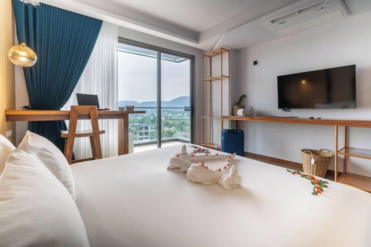 A hotel room with a bed, a desk with a laptop, a TV, and a view of the outdoors.