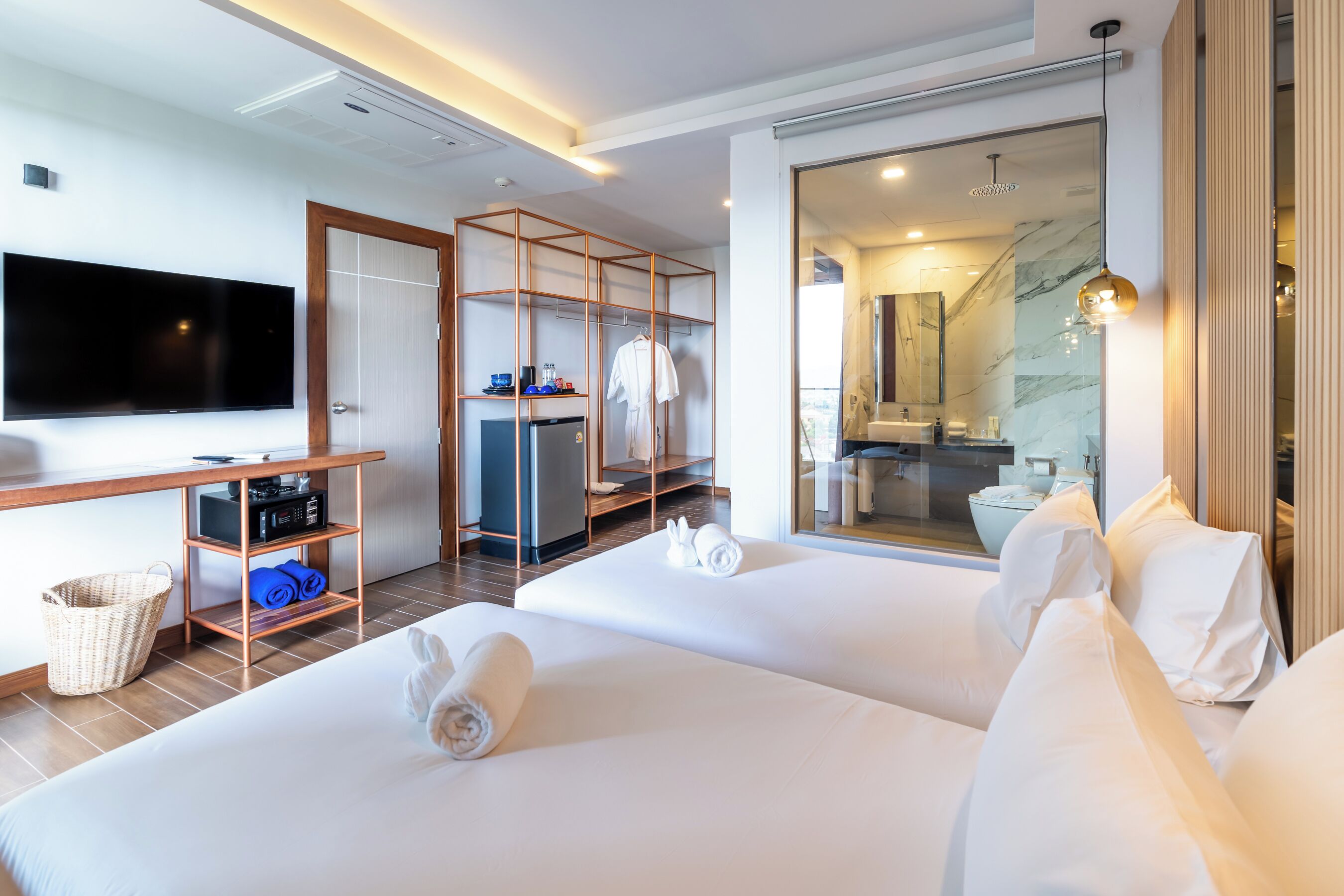 Ein Hotelzimmer mit zwei Betten, einem Fernseher, einem Schreibtisch, einem Kleiderschrank und einem durch eine Glastür einsehbaren Badezimmer.