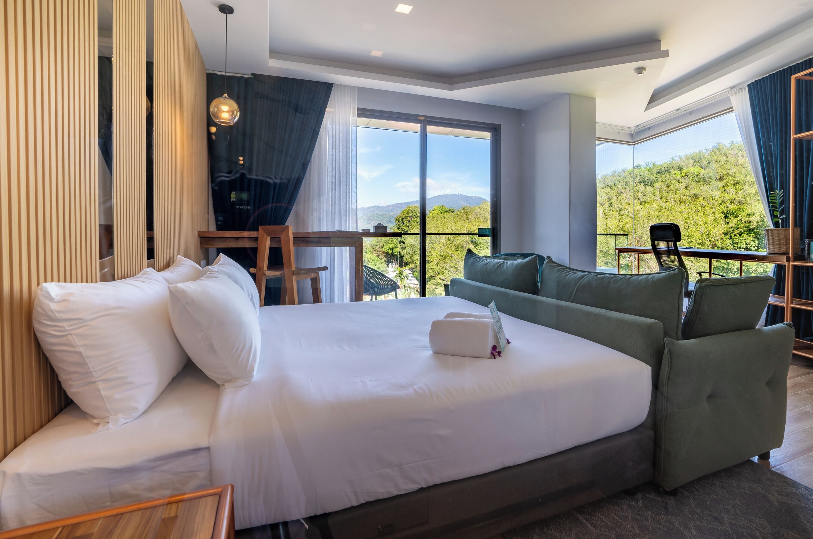 Een moderne hotelkamer met een groot bed, een groene bank, een eettafel en een balkon met uitzicht op de bergen.