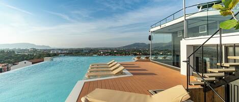 A rooftop pool area with lounge chairs, a wooden deck, and a glass balustrade overlooking a cityscape and mountains.