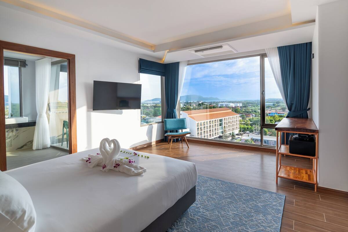 A hotel room with a large bed, a TV mounted on the wall, a view of the city, and a balcony with a chair.