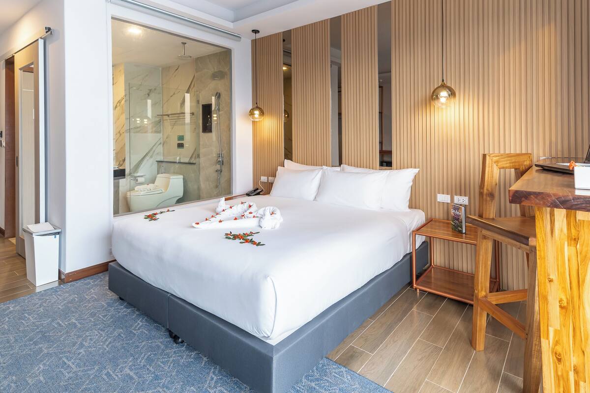 A modern hotel room with a large bed, a wooden desk, and a glass shower area.