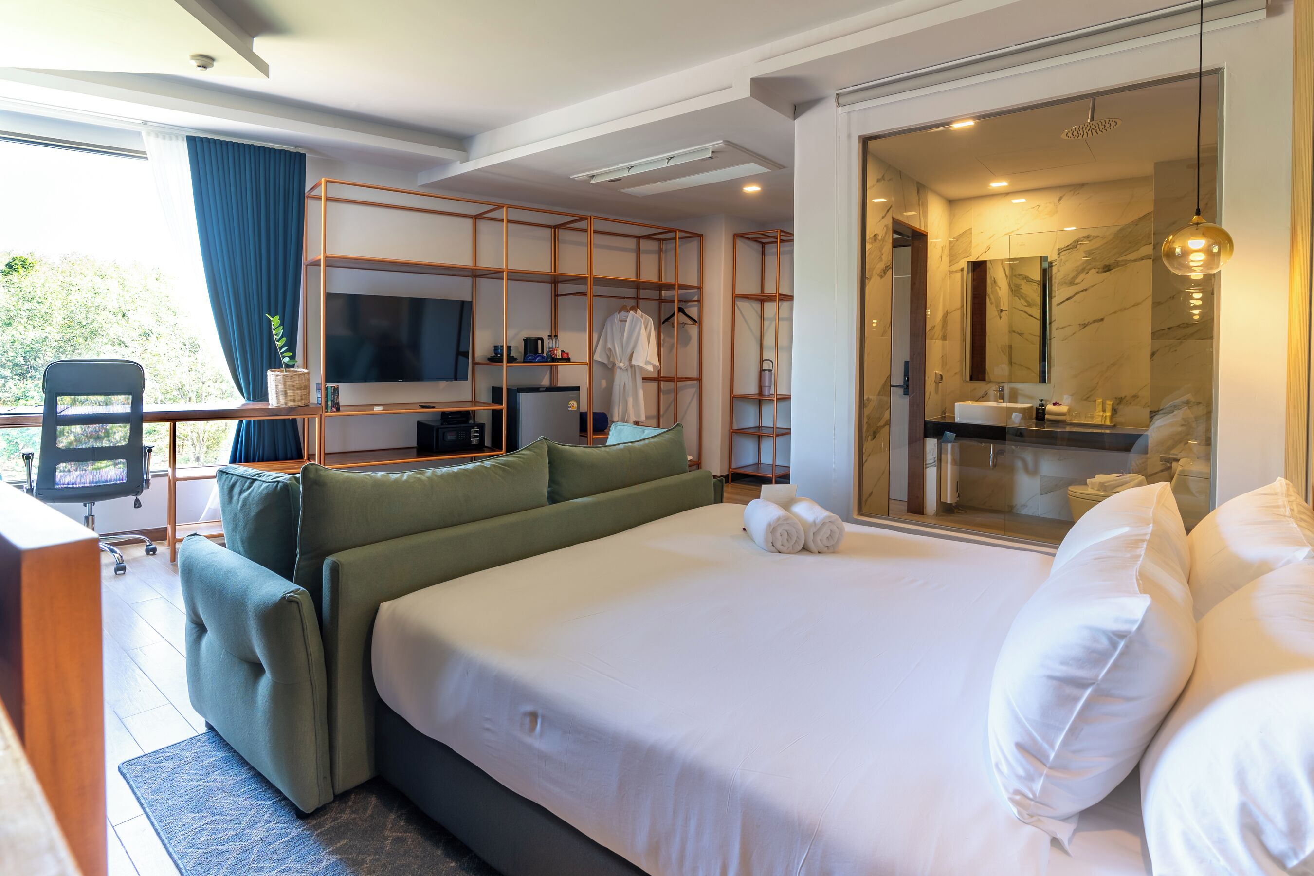 A modern hotel room with a large bed, a desk with a TV, a seating area, and a bathroom with a mirror and sink.