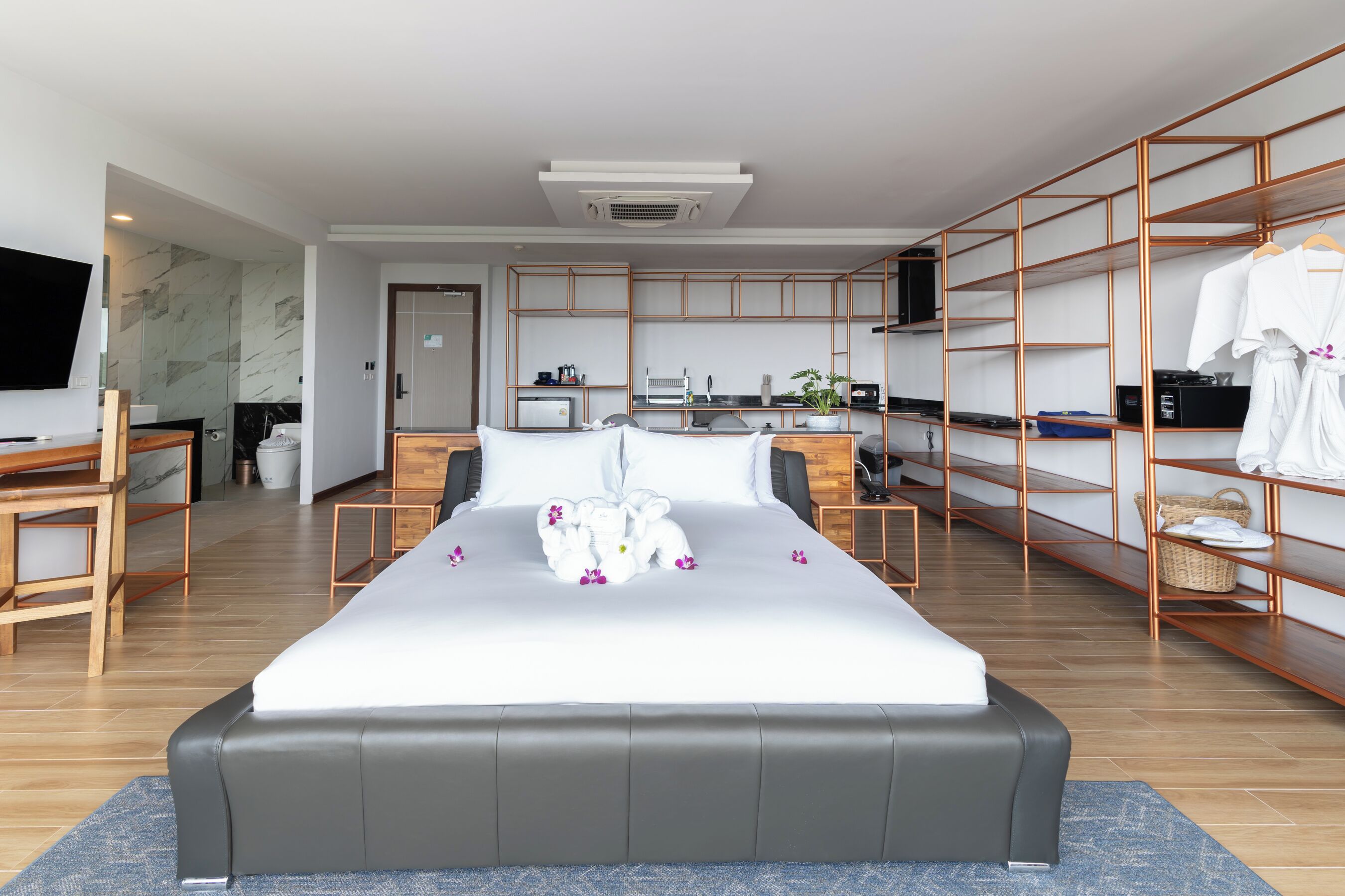 A modern hotel room with a large bed, a desk, a television, a kitchen area, and a wardrobe with shelves.