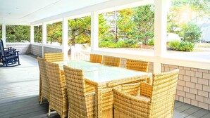 A covered patio with a glass-top table and wicker chairs, surrounded by white walls and large windows overlooking a garden.