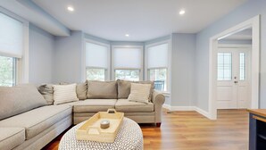 A modern living room with a sectional sofa, a round ottoman, and a window with blinds.