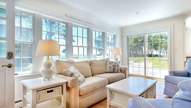 A spacious living room with a beige sofa, two side tables with lamps, and a view of the outdoors through large windows. - Seven Gables by Bayley Vacation Rentals (Kennebunk)