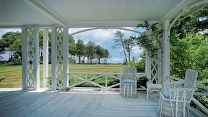 A covered porch with white lattice columns and a wooden deck, overlooking a grassy field and trees. - Seven Gables by Bayley Vacation Rentals (Kennebunk)