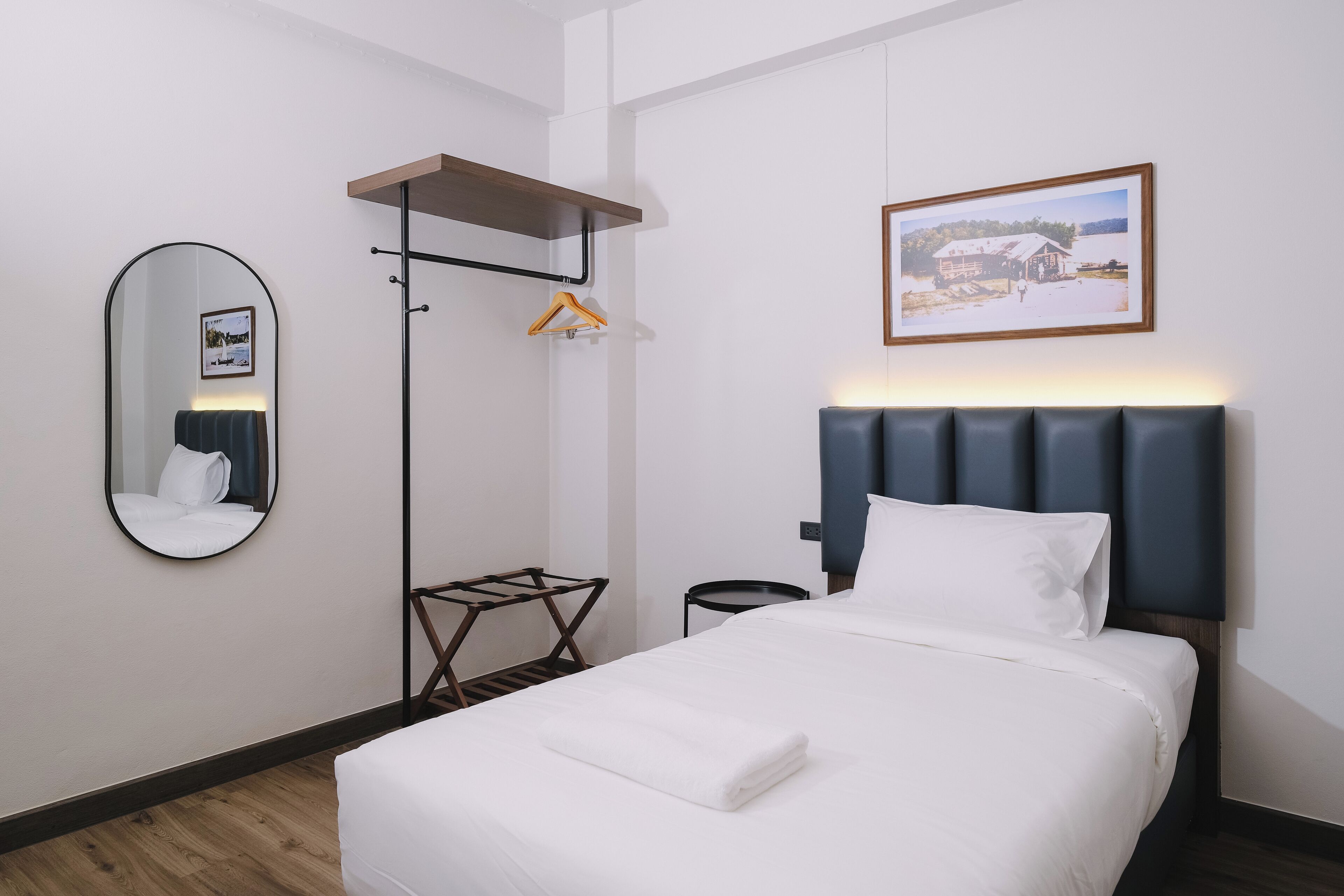A neatly arranged hotel room with a bed, a bedside table, a mirror, a hanging coat rack, and a framed picture on the wall.
