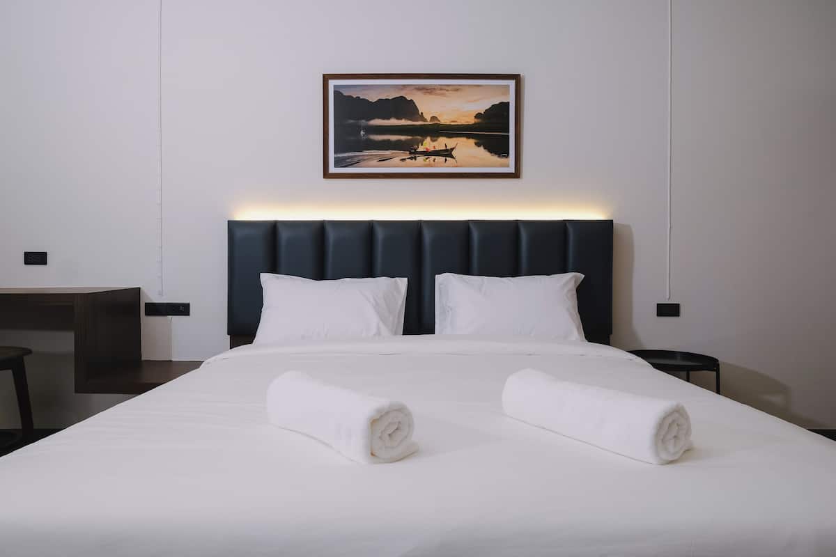 Un lit bien fait, avec deux serviettes enroulées, une tête de lit et un tableau représentant un paysage accroché au mur.