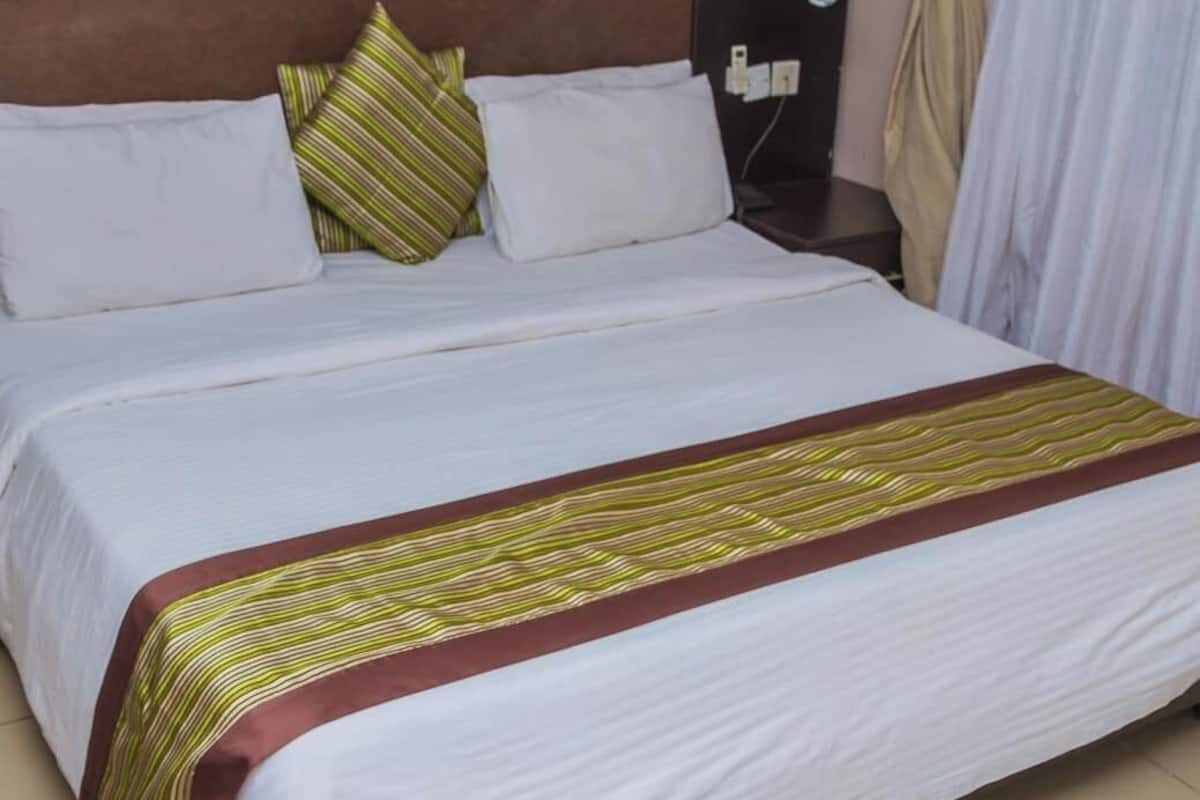 A neatly made bed with white linens and a striped blanket, a bedside table with a lamp, and a curtain partially drawn.