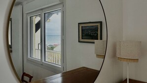 Un mirall rodó reflecteix una habitació amb una taula de fusta, una cadira, una finestra amb vista al mar i una làmpada sobre un tr ípode.