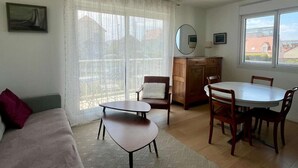 Un saló amb un sofà, una taula de centre, una taula de dinar amb cadires, un armari de fusta i una finestra gran amb vista a edificis.