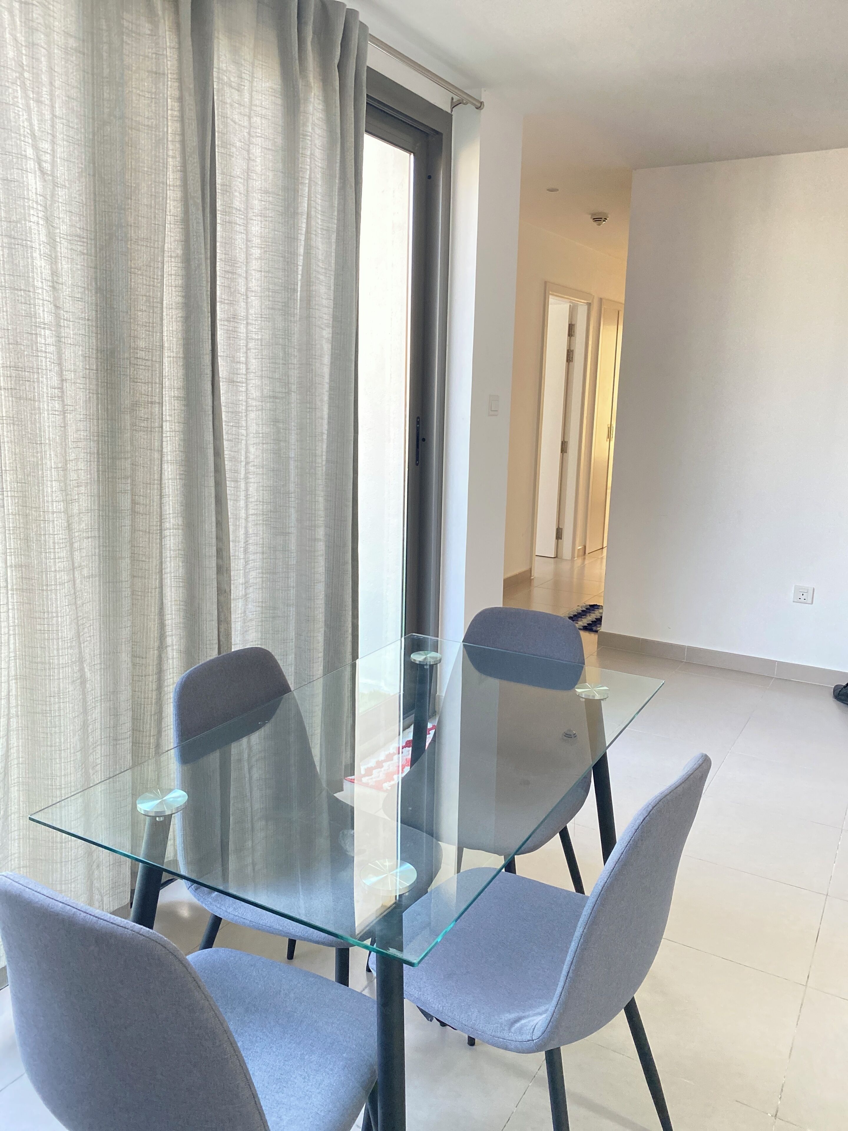 Un espace repas moderne avec une table en verre et quatre chaises grises, une fenêtre avec des rideaux et un mur blanc uni.