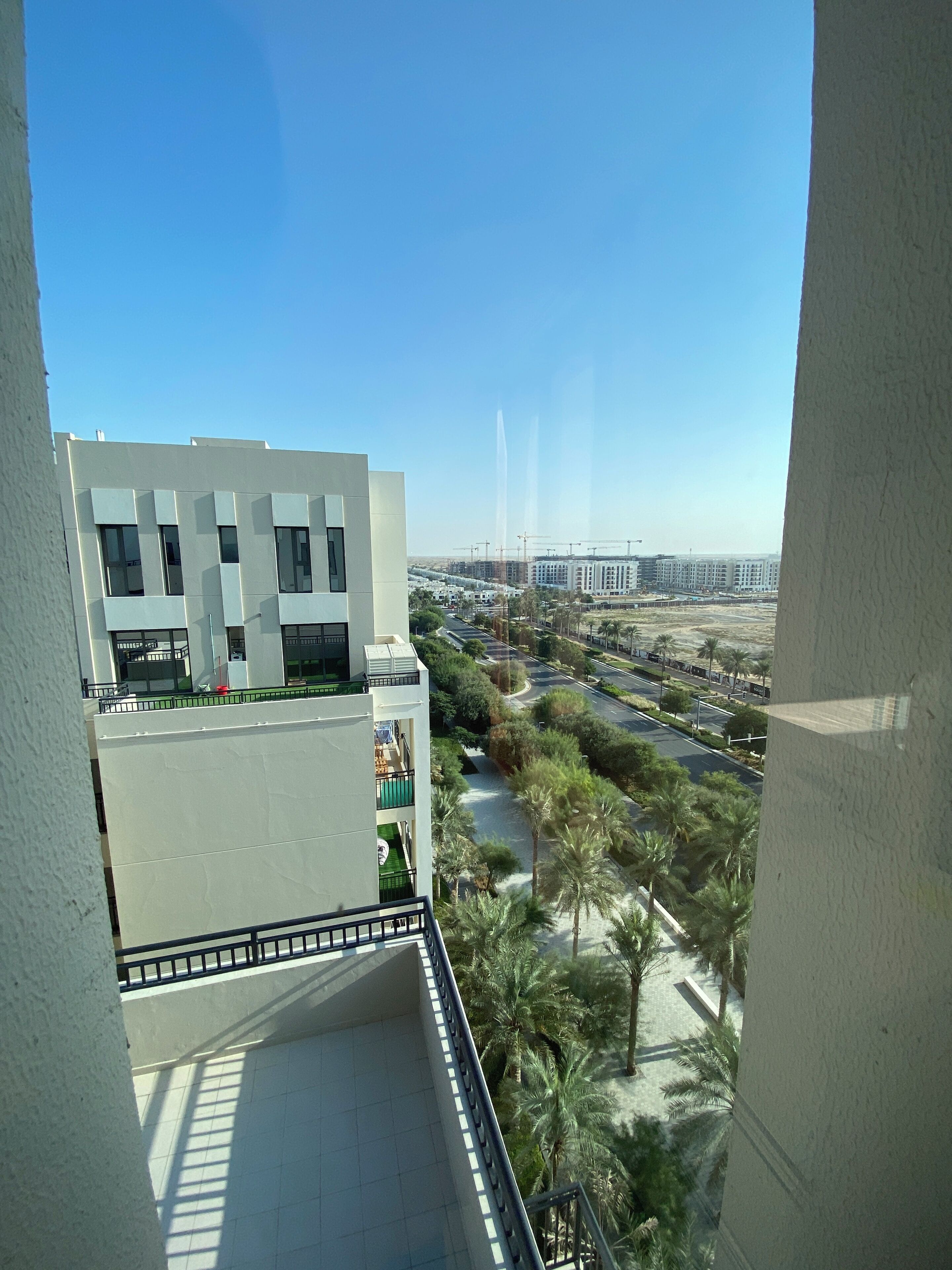A modern building with a balcony overlooking a landscaped area with palm trees and a road.